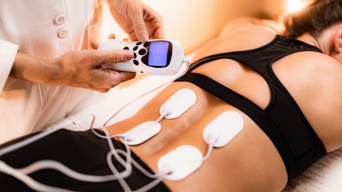 TENS kezelés során egy nő hátán elektródák találhatók, miközben a kezelő a készüléket állítja.