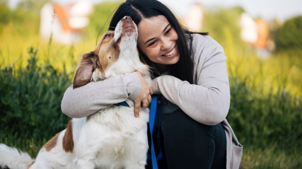 Egy nő boldogan öleli kutyáját a zöld fűben, szeretetteljes pillanatban.