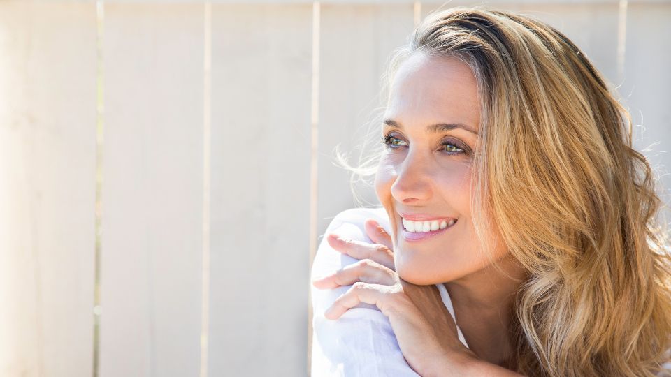 Egy boldog nő mosolyogva néz a kamerába, háttérben világos kerítés.